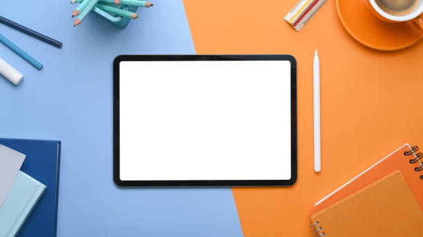 istock Overhead view of mock up digital tablet with blank screen surrounded by coffee cup, notebook and stationery on two tone blue and orange background. 1304655037