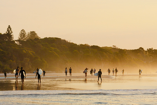 Horizontal landscape photo of a group of people, seen from a distance, walking on the beach, some of them carrying surfboards, as the afternoon sunset tints the beach, water and coastal forest with gold light. Byron Bay, east coast NSW.
