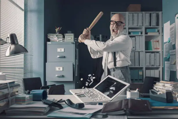 Crazy stressed businessman destroying his desk and laptop with a baseball bat, job burnout concept