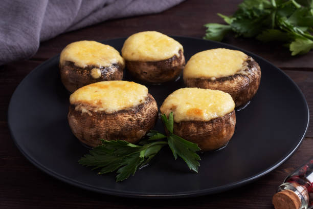 setas al horno rellenas de queso y hierbas en un plato negro. fondo de madera. - baked mushrooms fotografías e imágenes de stock