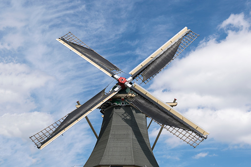 A windmill's striking silhouette against a soft blue sky, its blades adorned with the national colors. A proud Dutch flag flutters in the foreground, symbolizing national pride. The scene is framed with rustic wooden structures, possibly part of a mill complex, adding to the authentic atmosphere.