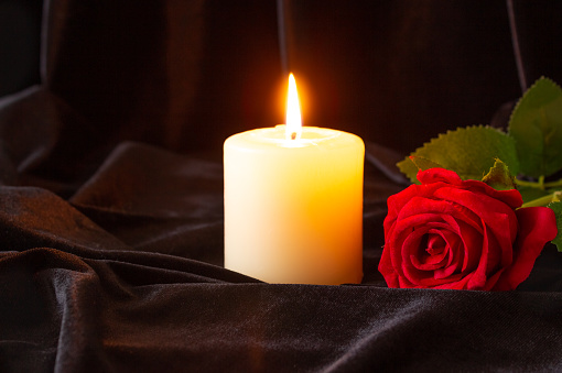A burning candle and a red rose on a black cloth background. The concept of condolence and religion. Focus in the background
