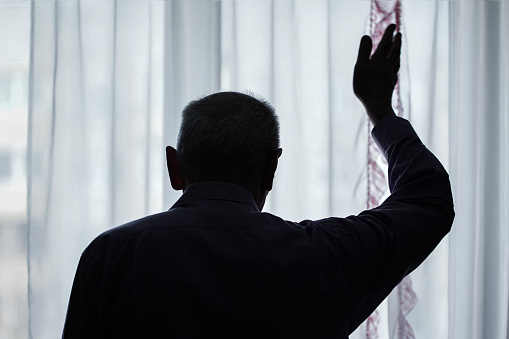 istock Dark silhouette of elderly man waving with hand goodbye gesture through window 1304693052