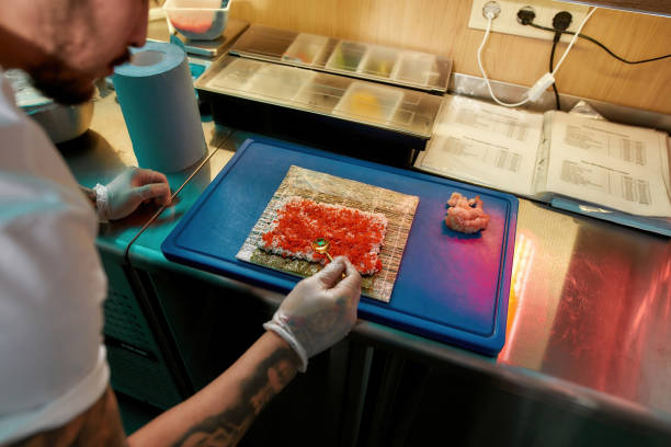 Hands in protective gloves of sushi chef spreading red caviar over cooked rice on seaweed wrap while making delicious rolls with shrimp at commercial kitchen Hands in protective gloves of sushi chef spreading red caviar over cooked rice on seaweed wrap while making delicious rolls with shrimp at commercial kitchen. Sushi making, Asian cuisine concept nori edible seaweed sheet stock pictures, royalty-free photos & images