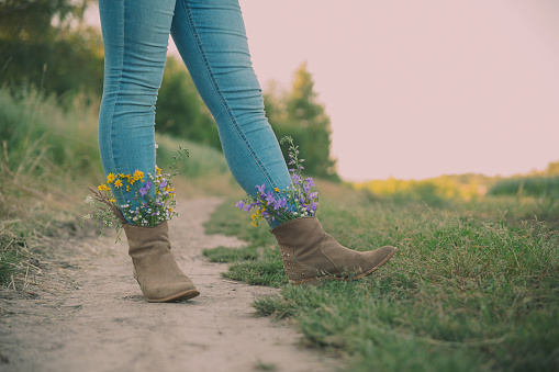 Female legs in jeans high boots with inserted flowers on the road in the field.