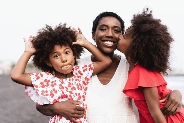 felici gemelle sorelle africane che baciano la madre sulla spiaggia - concentrati sul viso della madre - hair care immagine foto e immagini stock