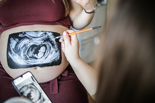 A young pregnant woman who is waiting for every moment of childbirth calmly poses and enjoys while the artist draws her belly. Happy pregnant woman. Body painting is in progress. The art of drawing on the body on the belly of a pregnant woman