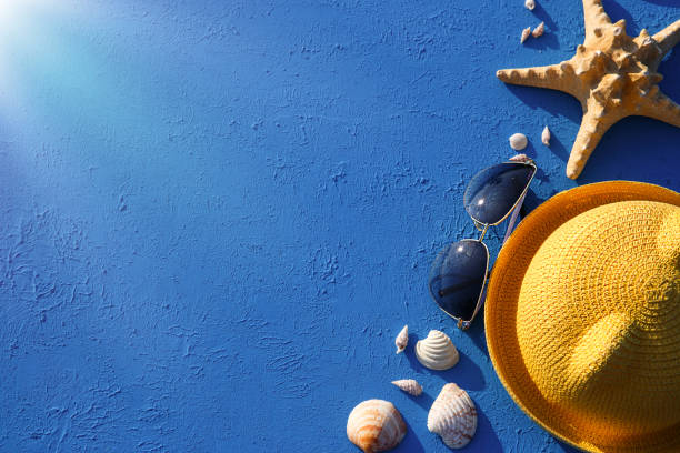 Frame with beach accessories on a nautical theme: yellow straw hat, sunglasses, starfish and shells on a blue background. Vacation concept, sea trip, UV protection, swimming. Copy space. Flatlay