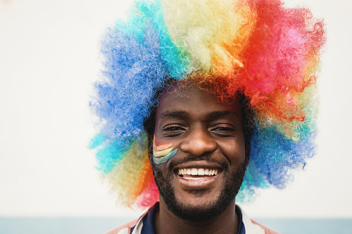 Happy young african man wearing lgbt rainbow flag wig - Focus on face