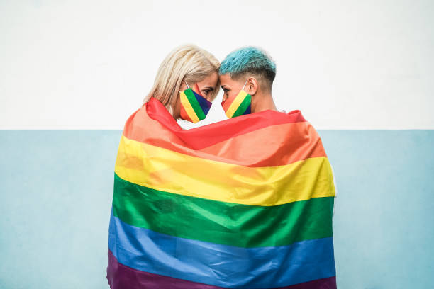 joven pareja de mujeres lesbianas bajo bandera arco iris lgbt usando máscaras en el desfile del orgullo gay - concéntrate en los rostros - homosexual rainbow gay pride flag flag fotografías e imágenes de stock