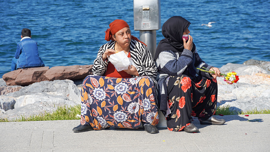 Two women sit on the pavement of seaside; one is resting , the other is eating her lunch in Konak, in Izmir, in Turkey on February 22, 2021