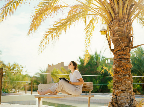 Young Caucasian woman sitting on the bench and writing in diary under the palm tree