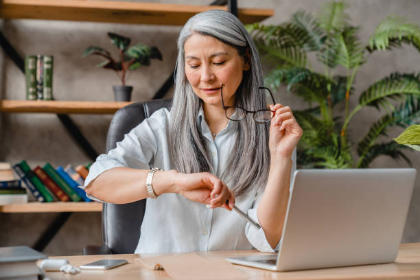 Busy mature businesswoman looking at her watch at her workplace in office Busy mature businesswoman looking at her watch at her workplace in office checking the time photos stock pictures, royalty-free photos & images
