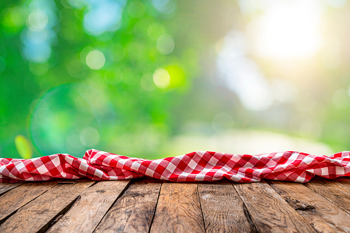 Spring backgrounds: eye view of an empty rustic wooden table with a crumpled red and white gingham cloth shot against defocused nature background. Useful copy space available for text, logo or product montage. Predominant colors are brown, green and yellow.