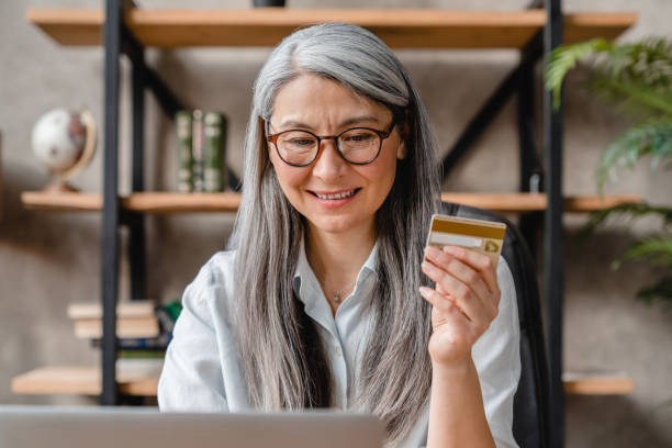 Cheerful senior grey-haired woman holding credit card for online shopping in office Cheerful senior grey-haired woman holding credit card for online shopping in office credit card paying banking business stock pictures, royalty-free photos & images