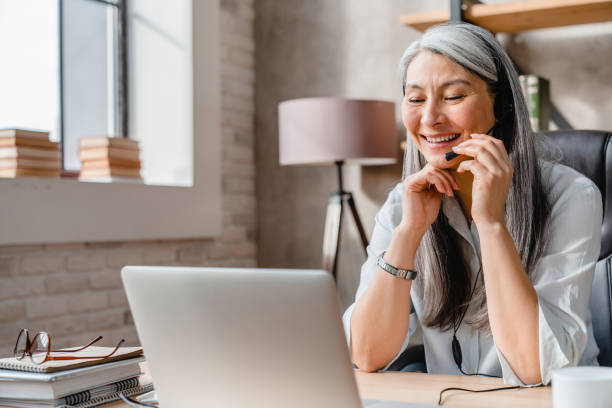 alegre mujer bastante madura trabajadora del centro de llamadas usando portátil en la oficina - women on the phone headset service fotografías e imágenes de stock