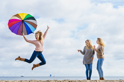 Female jumping with colorful umbrella her two friends are making fun of her laughing and looking.