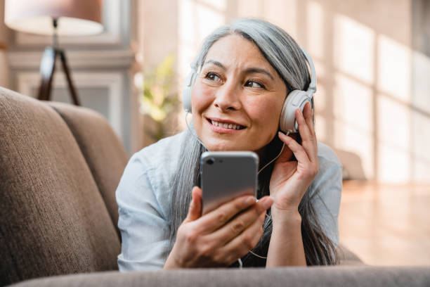 happy caucasian mature woman listening to the music on the phone on the couch - music listening women relaxation imagens e fotografias de stock