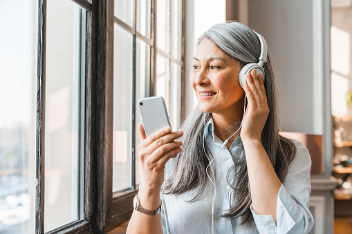 Excited mature grey-haired woman listening to the music on the phone at home