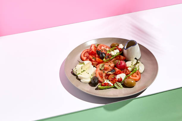 istock Tomato, olives and mozzarella salad . Restaurant appetizer plate on white table with pink wall. Day sunlight with hard shadow of fern palm leaves. Summer or spring restaurant food concept 1304729807