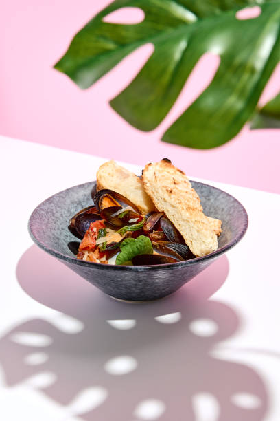 istock Mussel with bread restaurant plate. Restaurant appetizer plate on white table with pink wall. Day sunlight with hard shadow of monstera palm leaves. Summer or spring restaurant food concept. 1304730001