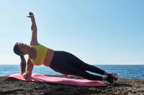 FITNESS WOMAN IN SPORTS SET TRAINING WITH ELASTIC BAND, WEIGHTS, GYM EXERCISES, IN FRONT OF THE WATER.