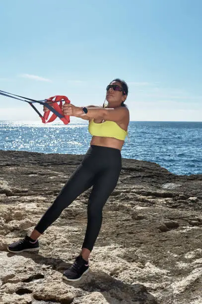 FITNESS WOMAN IN SPORTS SET TRAINING WITH ELASTIC BAND, WEIGHTS, GYM EXERCISES, IN FRONT OF THE WATER.