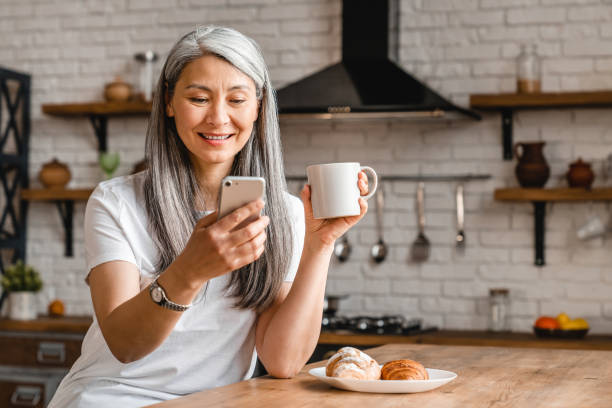 donna felice di mezza età che usa lo smartphone durante la colazione in cucina - smart phone asian ethnicity mobile phone women foto e immagini stock