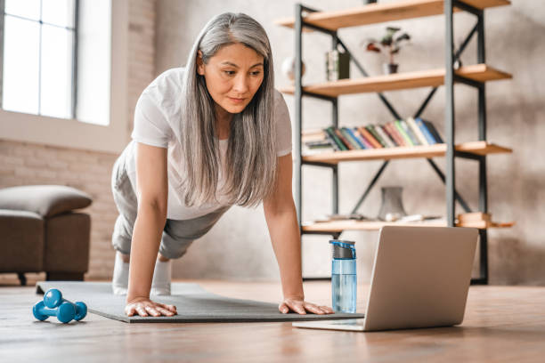 sportowa kaukaska kobieta w średnim wieku stojąca w pozycji deski za pomocą laptopa do treningu w domu - healthy lifestyle yoga vitality flexibility zdjęcia i obrazy z banku zdjęć