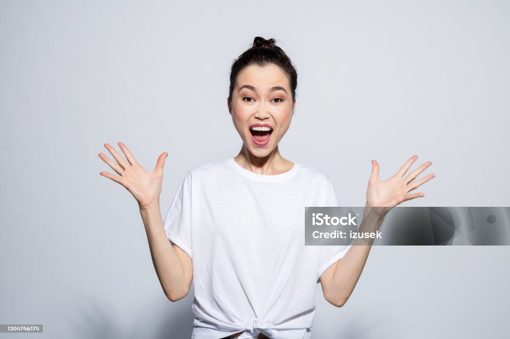 Excited young woman screaming at camera Portrait of happy, surprised asian young woman wearing white t-shirt, screaming at camera with raised hands. Studio shot, grey background. Women Stock Photo