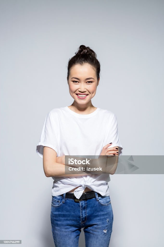 Friendly asian young woman standing with arms crossed Portrait of beautiful asian young woman wearing white t-shirt and jeans, looking at camera with arms crossed. Studio shot, grey background. Fashion Model Stock Photo