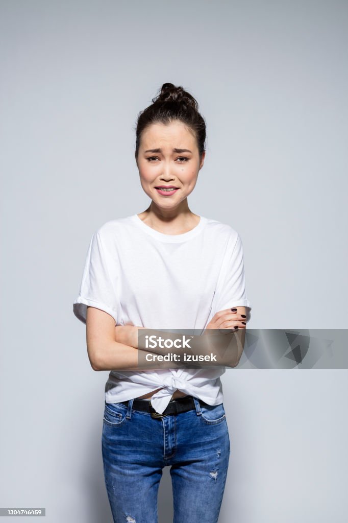 Friendly asian young woman standing with arms crossed Portrait of beautiful asian young woman wearing white t-shirt and jeans, laughing at camera with arms crossed. Studio shot, grey background. Fashion Model Stock Photo