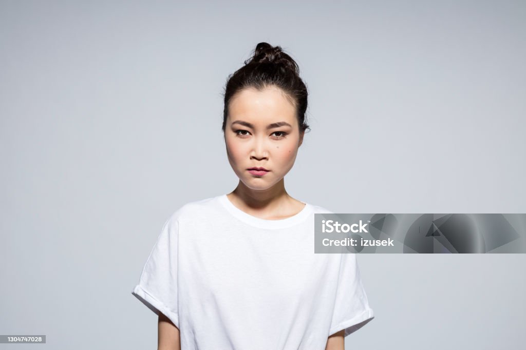 Headshot of angry asian young woman Portrait of frustrated asian young woman wearing white t-shirt, staring at camera. Studio shot, grey background. Portrait Stock Photo