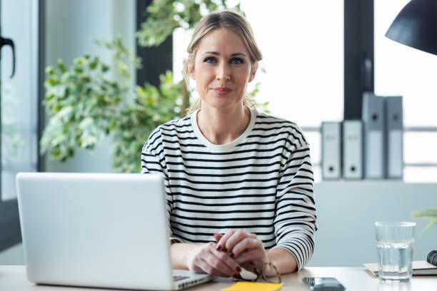 istock Beautiful mature business woman working with computer while looking at camera in the office at home. 1304747520