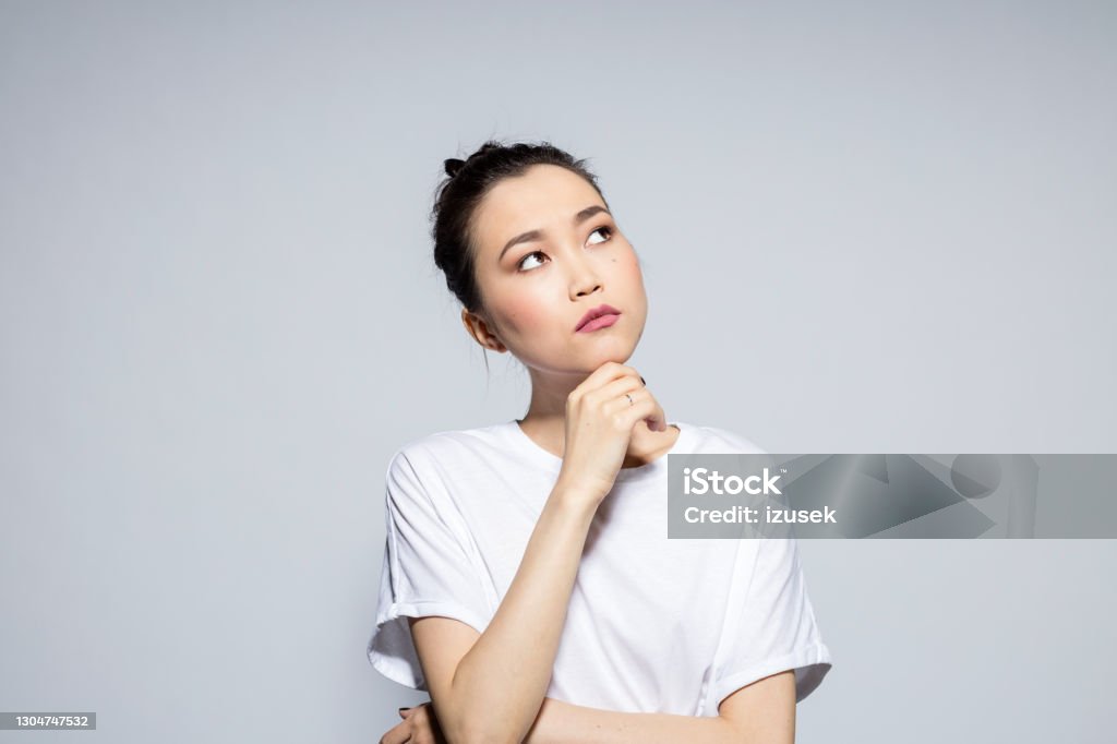 Pensive beautiful asian young woman Portrait of beautiful asian young woman wearing white t-shirt, looking away with hand on chin. Studio shot, grey background. Contemplation Stock Photo