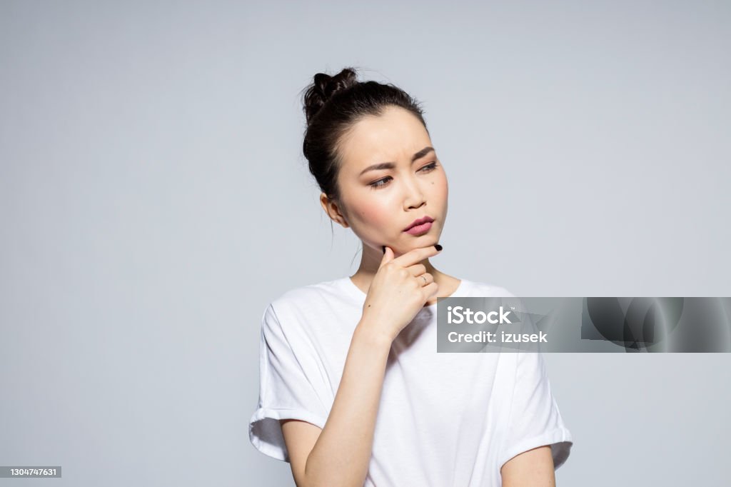 Pensive beautiful asian young woman Portrait of beautiful asian young woman wearing white t-shirt, looking away with hand on chin. Studio shot, grey background. 20-24 Years Stock Photo