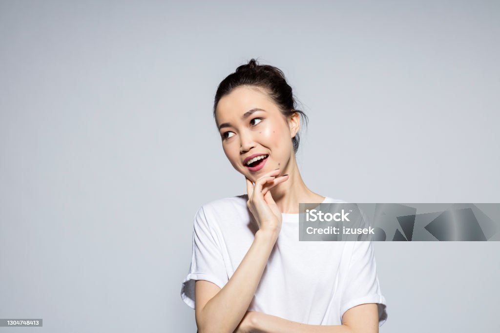 Portrait of surprised young woman Cheerful, beautiful asian young woman wearing white t-shirt, laughing with hand on chin, looking away. Studio shot, grey background. Women Stock Photo