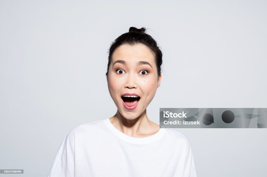 Surprised young woman staring at camera Headshot of happy, beautiful asian young woman wearing white t-shirt, staring and mouth open. Studio shot, grey background. Shock Stock Photo