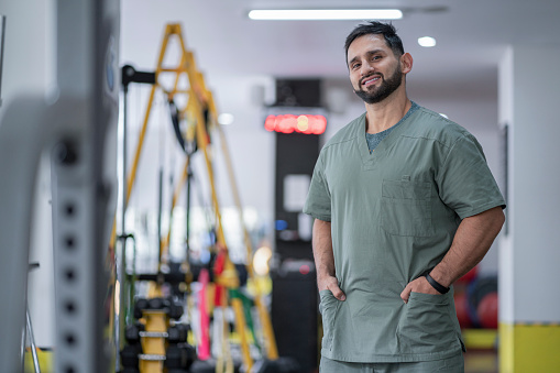Latin man of average age of 30 years with a black beard from the city of Bogota, a physiotherapist, is standing inside the gym where he works dressed in his green antifluidic uniform looking at the camera that portrays him