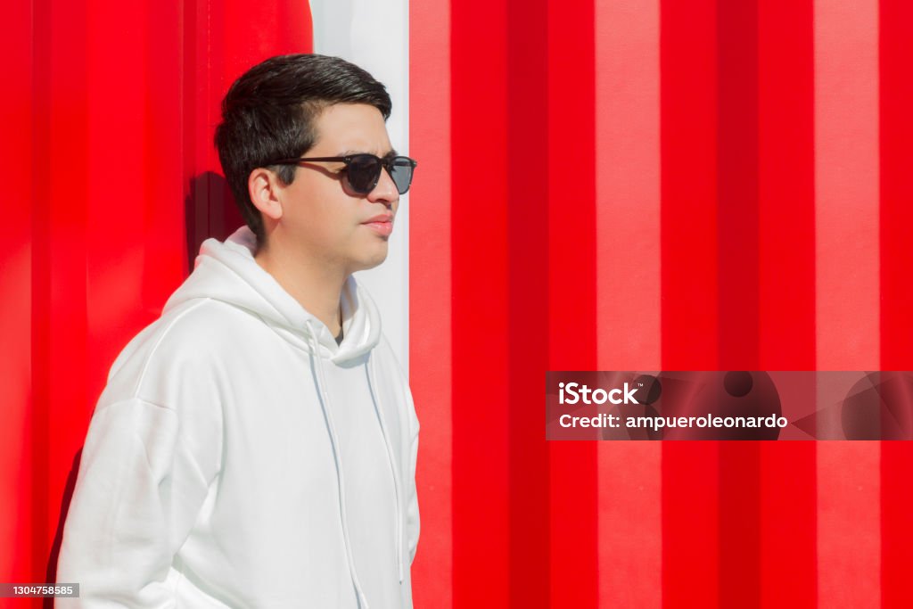 Young latin ethnic stylish man Portrait in Miami Beach, Florida, USA Cheerful Young stylish ethnic smiling hipster hippie indie Art Graphic Designer Publisher latin man portrait at the street in Miami Beach, Florida, USA. He's wearing a nice fashionable black sunglasses and a pure clean white sweater, on back, a colorful red background in a sunny day. Males Stock Photo