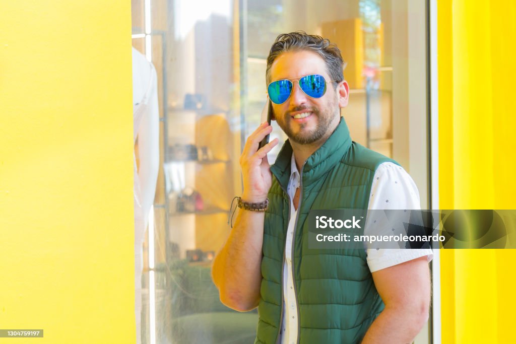 Young latin stylish man texting by cellphone in Miami Beach, Florida, USA Cheerful Young stylish smiling hipster hippie indie Art Graphic Designer Publisher latin man taking by cellphone at the street in Miami Beach, Florida, USA. He's wearing a nice fashionable blue sunglasses, green winter jacket, a white shirt and a red jeans pants, on back, a colorful yellow background in a sunny day. Adult Stock Photo