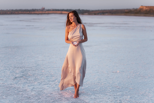 Young blonde woman in evening airy pastel pink powdery dress, holds pieces of crystallized white salt in hands. Girl with natural make-up, hair developing. Salt mining trip, walking on water at sunset