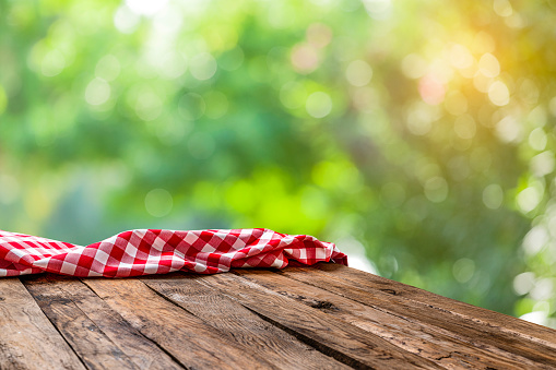 Spring backgrounds: eye view of an empty rustic wooden table with a crumpled red and white gingham cloth shot against defocused nature background. Useful copy space available for text, logo or product montage. Predominant colors are brown, green and yellow.