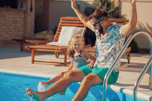 Beauitful cheerful father and daughter sitting at the edge of a swimming pool and having fun outdoors on a hot sunny summer day