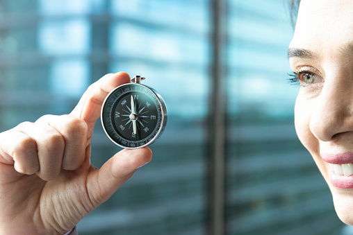 Businesswoman is holding a compass in hand in front of a skyscraper and looking at compass with a warm smile.