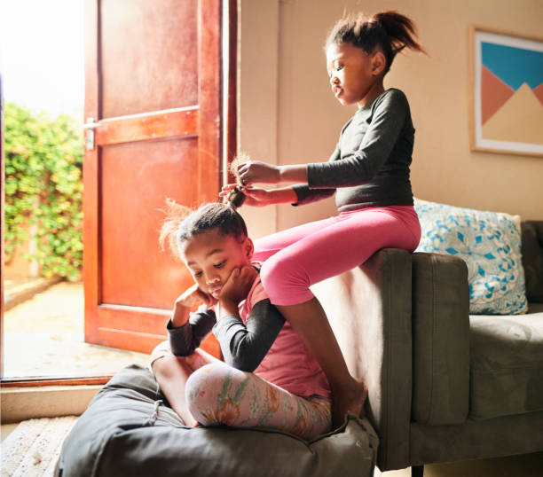 Cute little girl doing her sister's hair at home Little girl putting her sister's hair in pigtails while sitting together in their living room at home little black girl hairstyle stock pictures, royalty-free photos & images