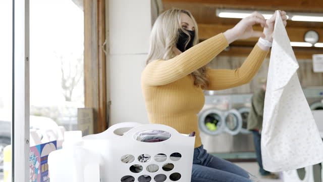 Millennial Woman Wearing Protective Face Mask Folding Laundry at Laundromat