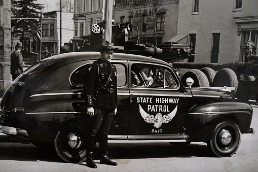 Lima, Ohio, USA - July 20, 1940: Ohio state highway patrolman standing by patrol car, military equipment going through Ohio town, black and white