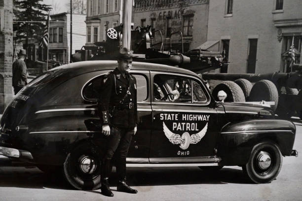 street scene from 1940, military equipment, Ohio state highway patrol Lima, Ohio, USA - July 20, 1940: Ohio state highway patrolman standing by patrol car, military equipment going through Ohio town, black and white ohio photos stock pictures, royalty-free photos & images