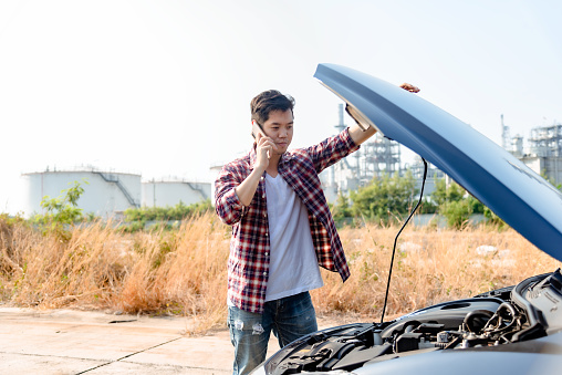 Asian man is talking on the phone for help due to a broken car during the trip. A man talking on a mobile phone in front of a car hood.